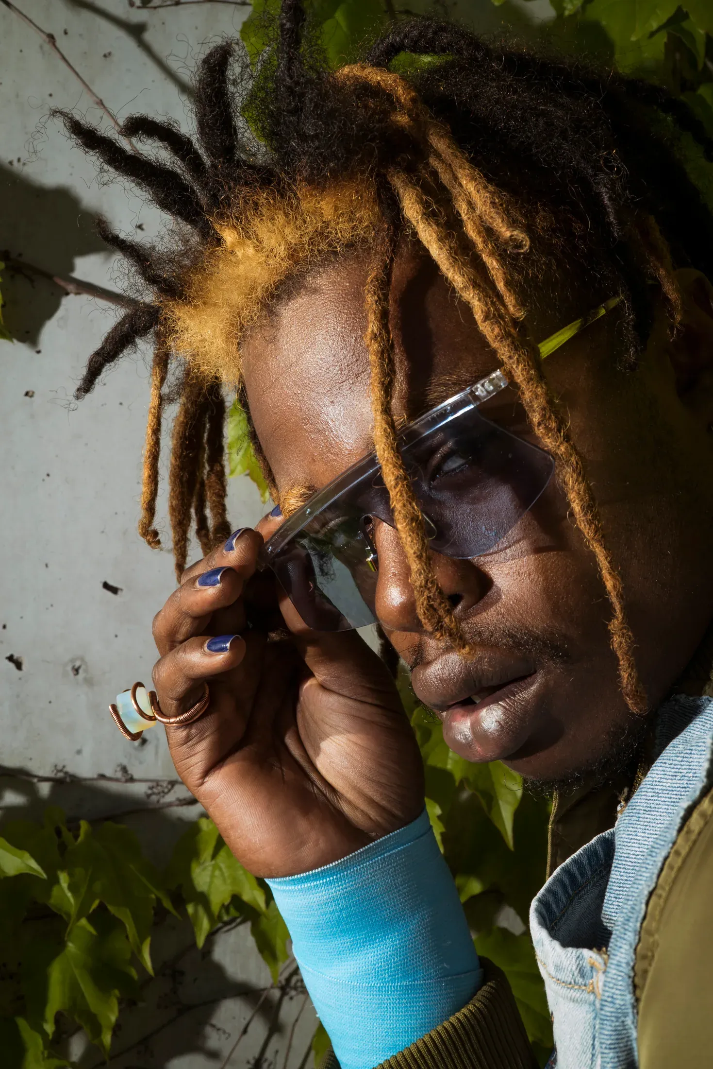 Close portrait of a person adjusting sunglasses beside green leaves.