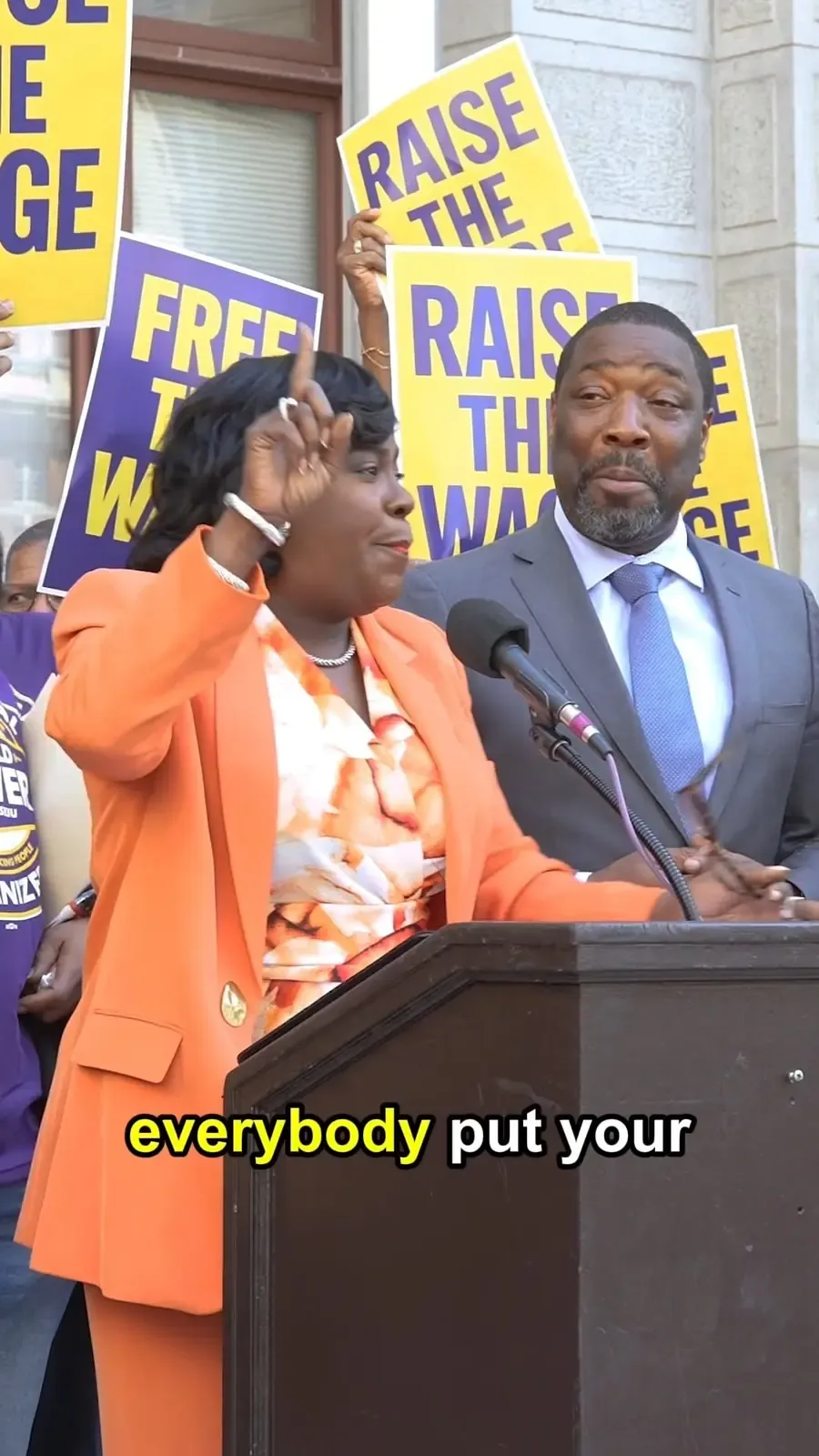 A speaker stands before Raise the Wage signs during a Philadelphia minimum wage campaign action.